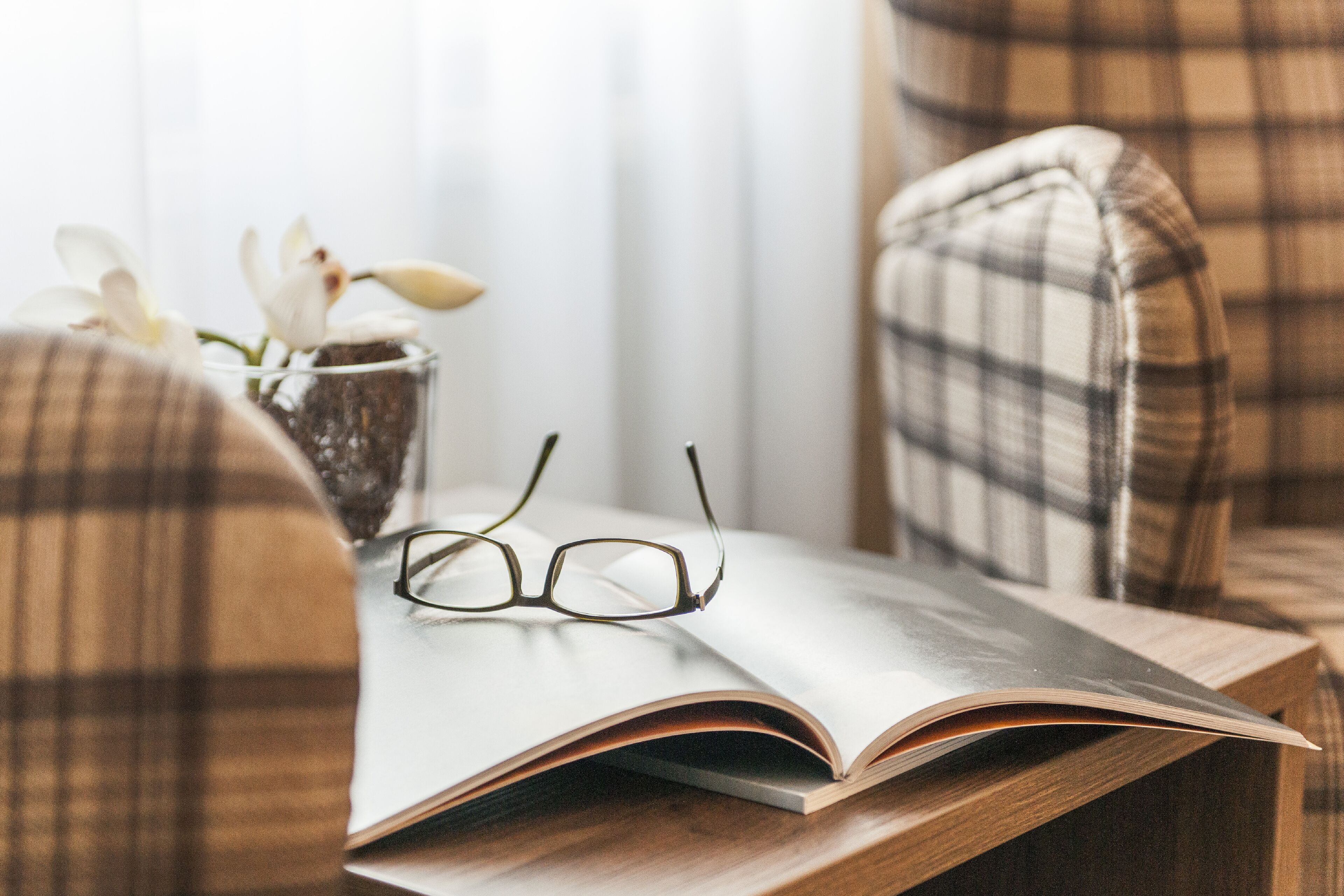 A plaid chair, a book, and a pair of glasses on a wooden table.
