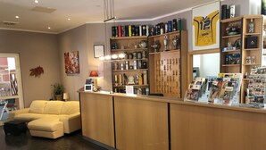 A reception area with a wooden counter, a sofa, and a wall decorated with various items including a framed certificate, a lamp, and a shelf with books and trophies. - Brandiser Parkhotel (Brandis)