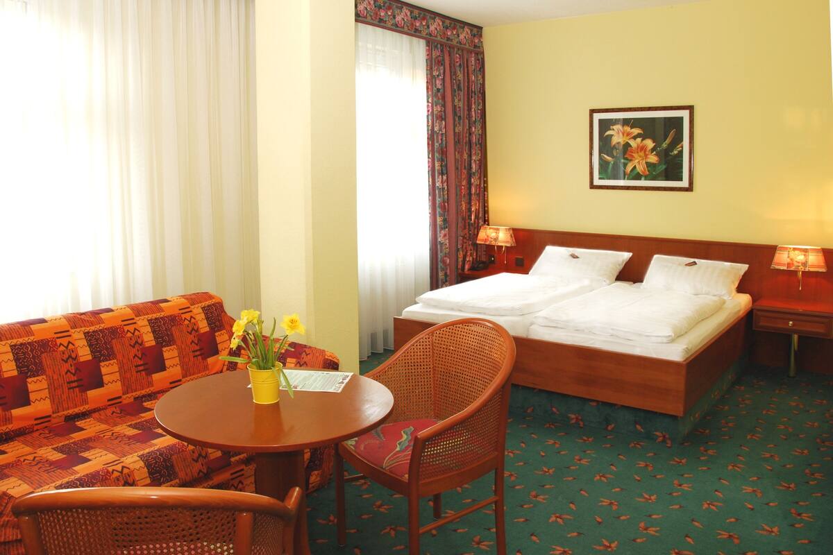 A hotel room with a bed, a sofa, a round table with a vase of flowers, and a chair.