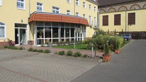 A yellow building with a red-tiled roof, large windows, and a small garden area with potted plants.
