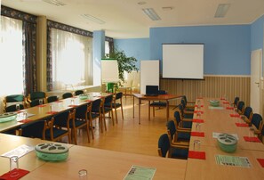 A conference room with long tables, chairs, a projector screen, and a large window with curtains.