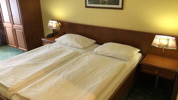 A double bed with white linens, two bedside tables with lamps, a wooden headboard, a framed floral picture on the wall, and a green carpet with leaf patterns.