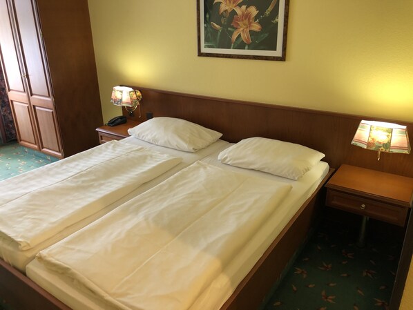 Una cama doble con sábanas blancas, dos mesitas de noche con lámparas, un cabecero de madera, un cuadro con motivos florales en la pared y una alfombra verde con diseños de hojas.