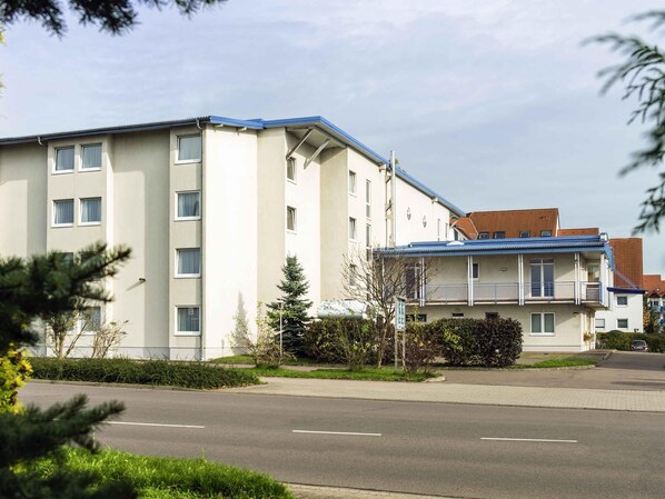 Un edificio residencial de varios pisos con techo plano, balcón y zona de estacionamiento al frente.