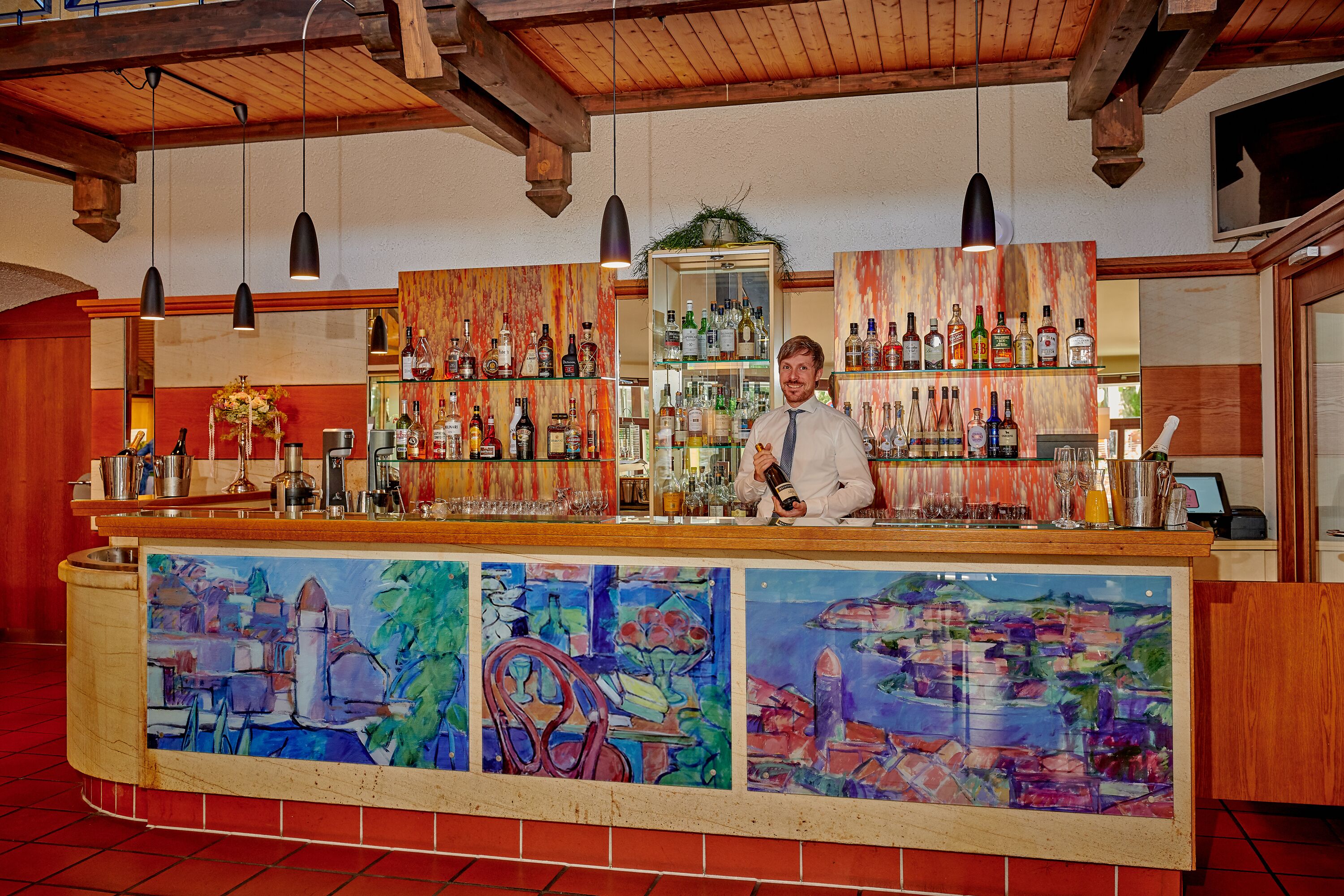 Eine Bar mit Theke, einem Barkeeper, Regalen voller Spirituosenflaschen und einem farbenfrohen Wandbild.