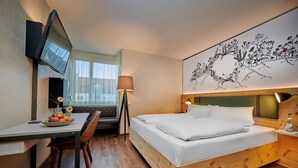 Habitación de hotel con cama, escritorio con silla, lámpara, televisor montado en la pared y una obra de arte en la pared.