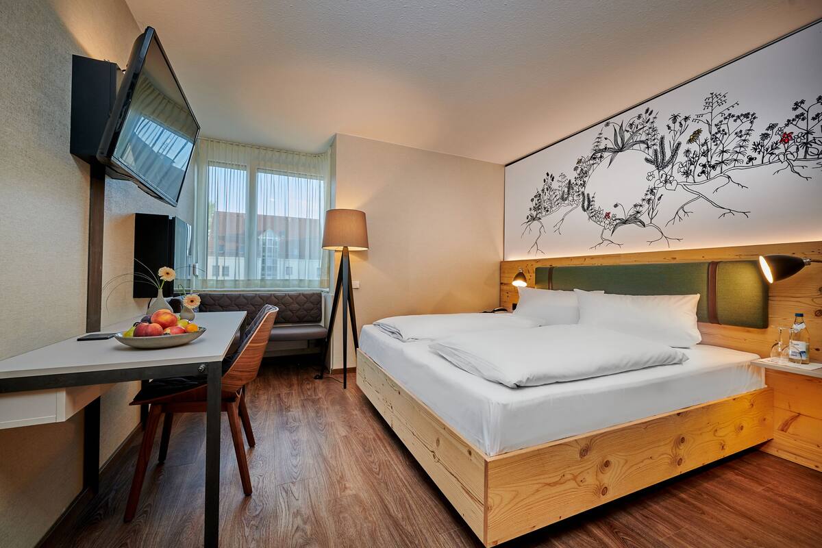 Een hotelkamer met een bed, een bureau met stoel, een lamp, een aan de muur bevestigde televisie en een kunstwerk aan de wand.