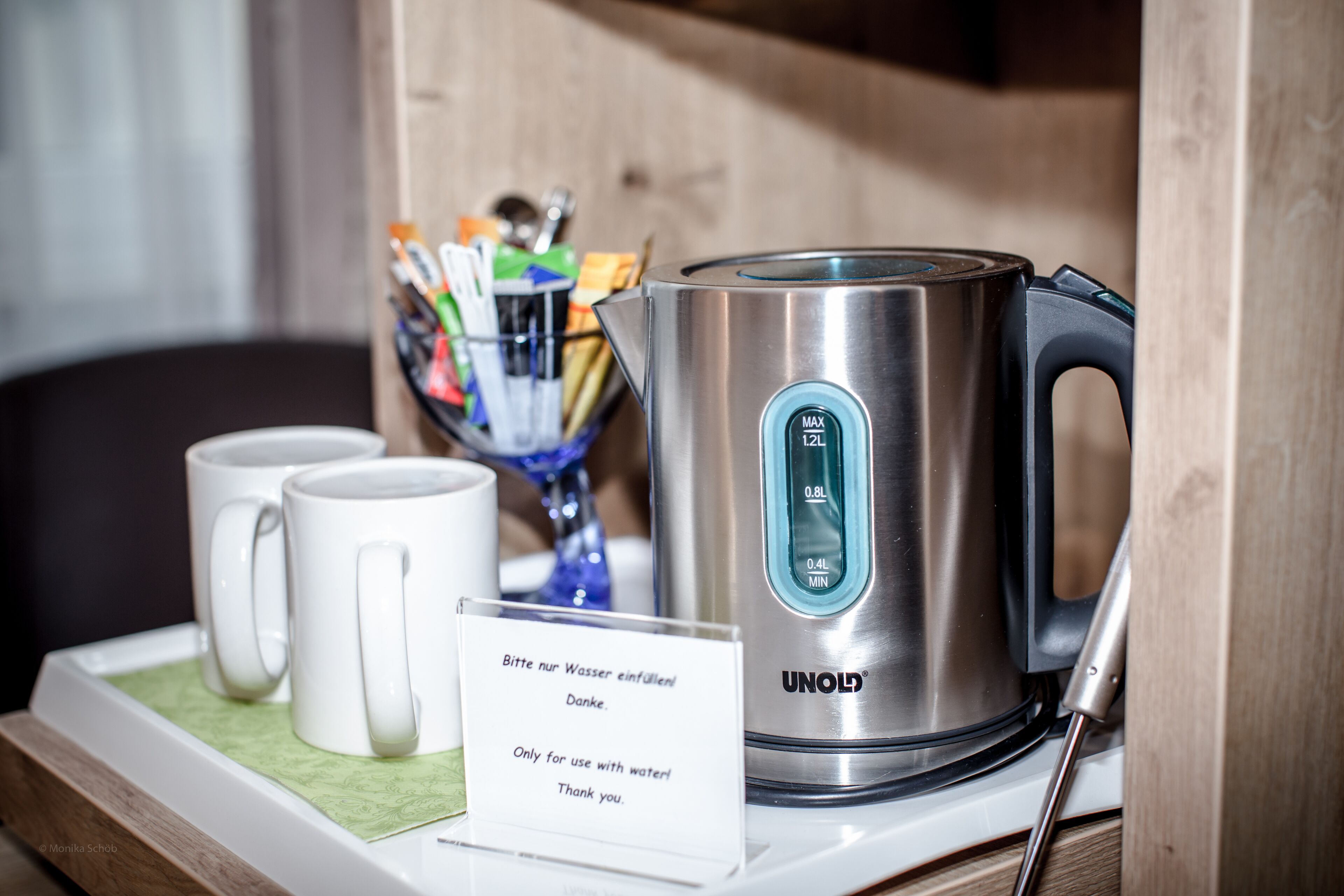 A stainless steel kettle with a digital display, two white mugs, and a sign with instructions in German.