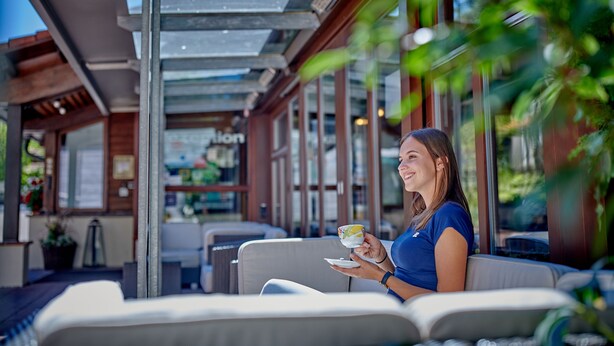 Una mujer sentada en un banco acolchado al aire libre, sosteniendo una taza y un platillo.