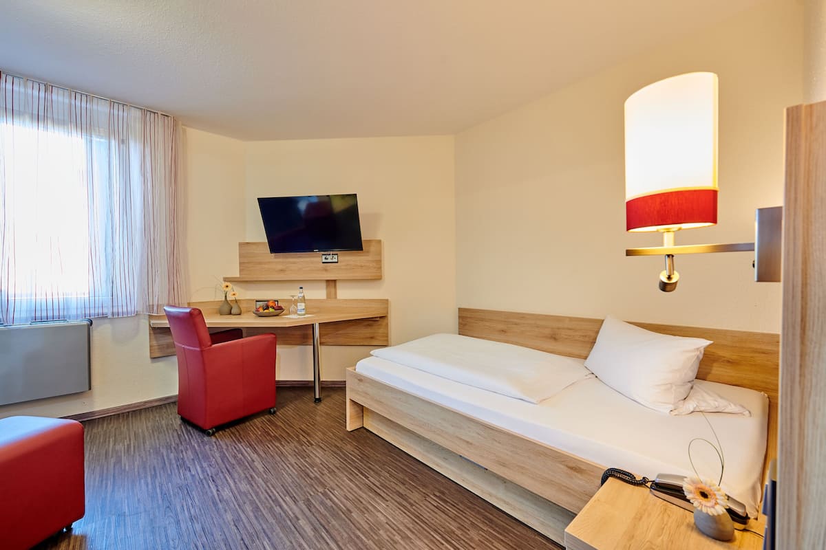 Een hotelkamer met een bed, een bureau met een televisie, een rode stoel, een lamp en een klein tafeltje met een vaas en een bloem.