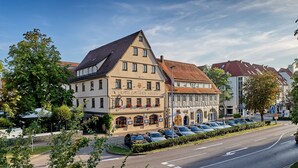 Un hotel de estilo arquitectónico europeo tradicional, con un techo a dos aguas destacado y una fila de coches aparcados frente a él.