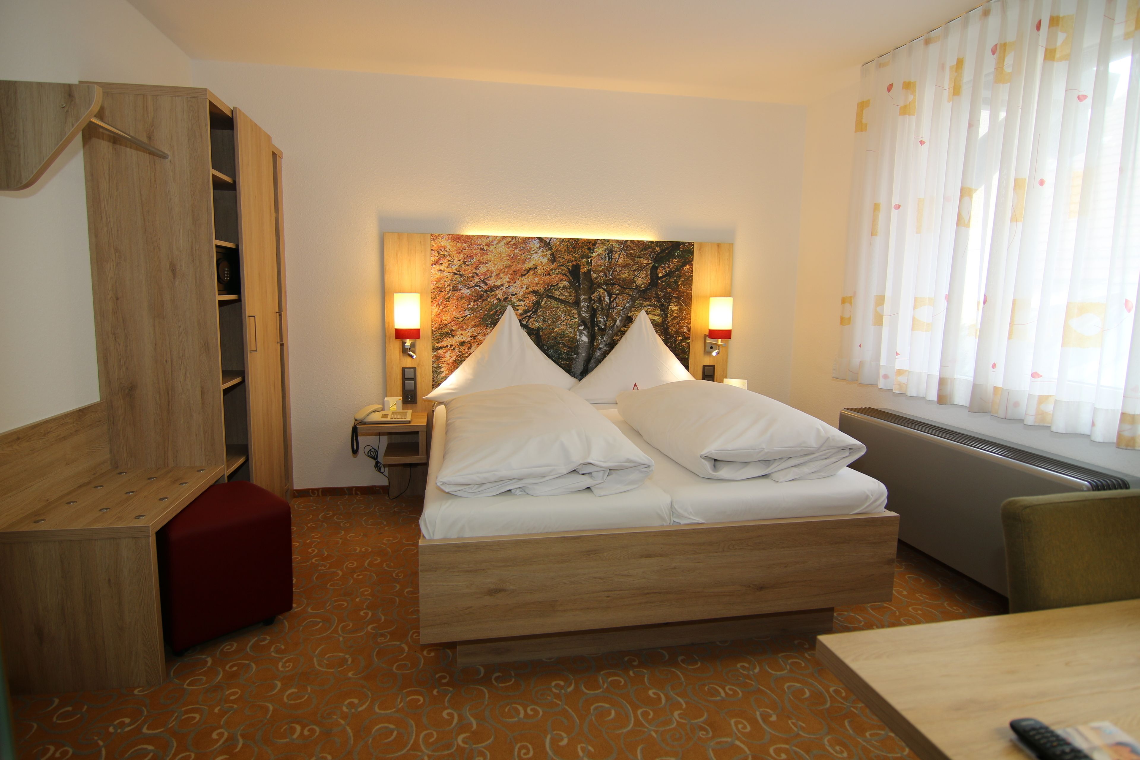 Ein Hotelzimmer mit Bett, Nachttischlampen, einem Holzschrank, einem roten Hocker und einem Teppich mit Muster.
