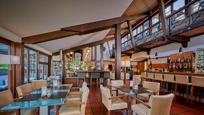 Un restaurante con mesas y sillas de vidrio, una zona de bar con estantes llenos de botellas y ventanas amplias que dejan entrar luz natural.