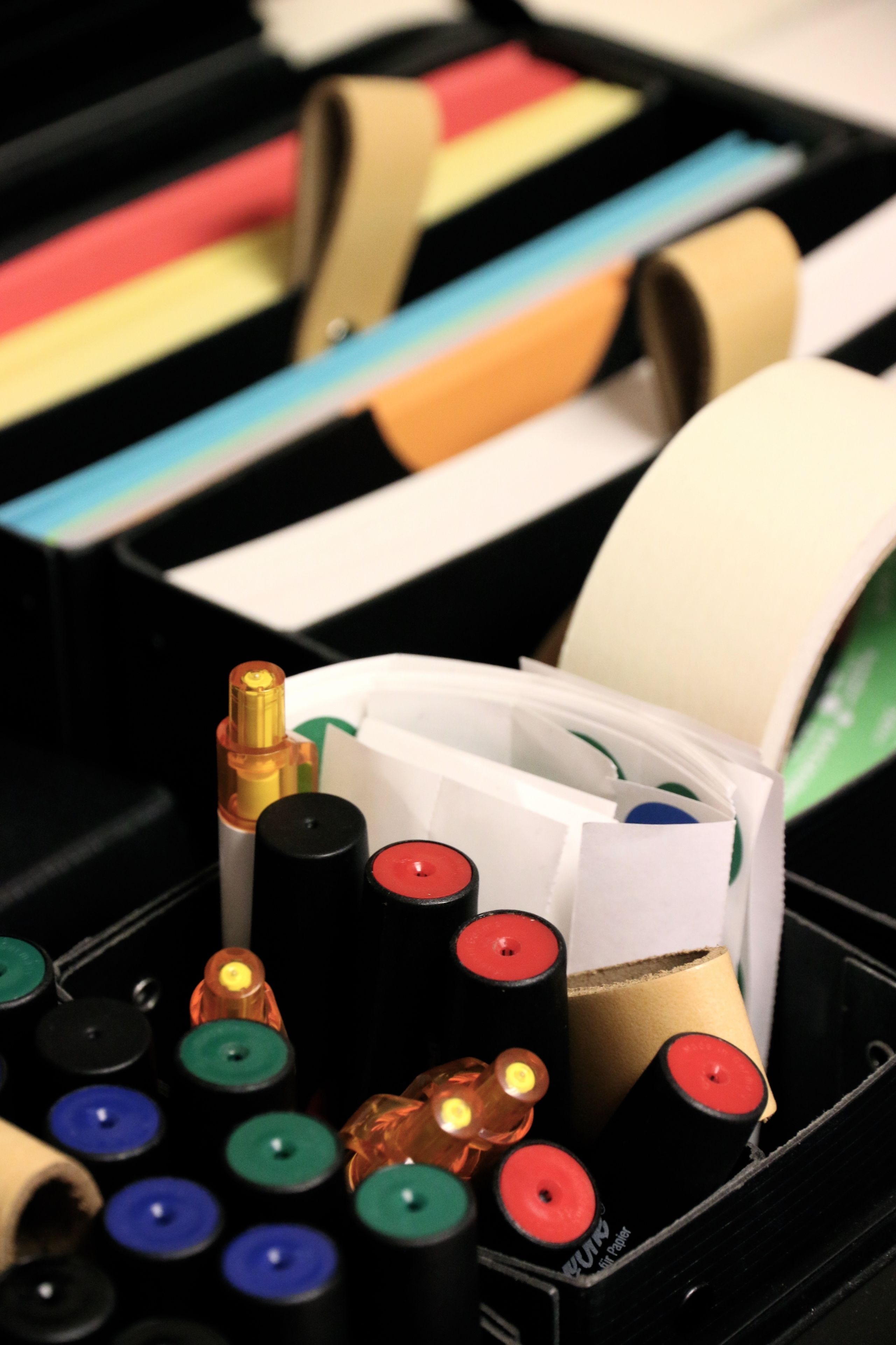 A black storage box containing various office supplies including colored markers, tape, and a roll of paper.