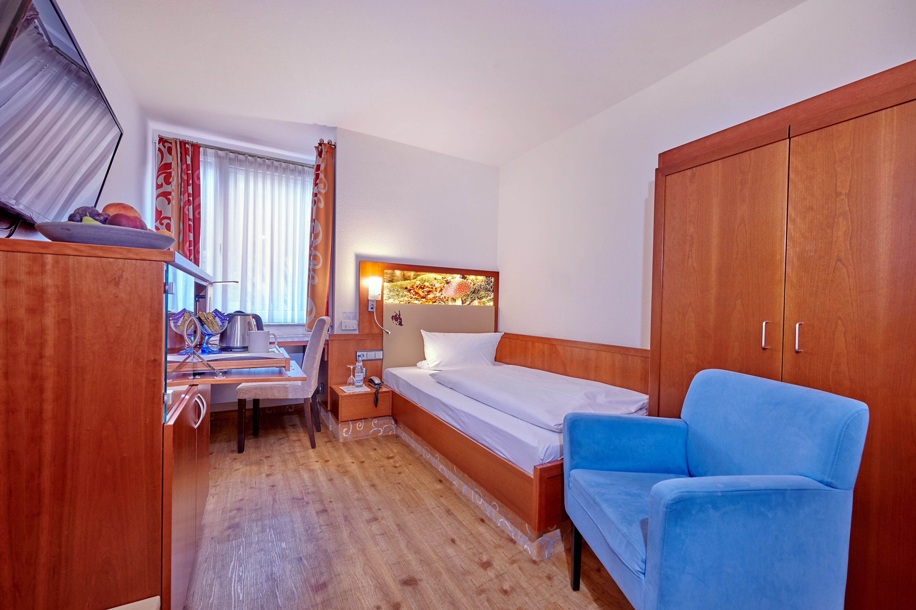 A hotel room with a wooden wardrobe, a bed with white linens, a blue armchair, a desk with a chair, and a window with curtains.