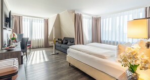 A hotel room with a bed, a desk with a computer, a sofa, and a dining table with a vase of flowers. - PLAZA Hotel Buchhorner Hof (Friedrichshafen)