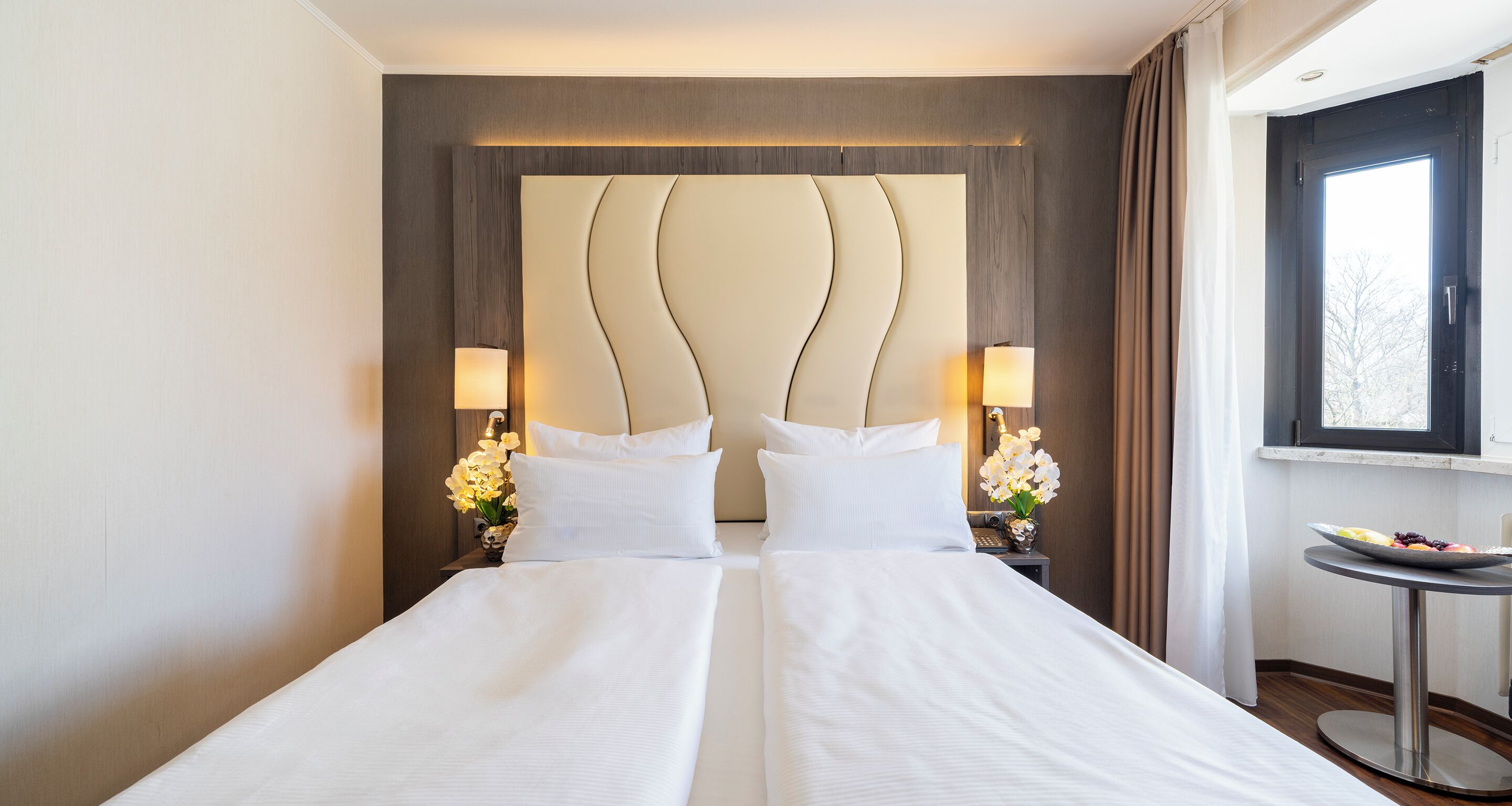 Quarto de hotel com uma cama grande, duas mesas de cabeceira com abajures, uma mesinha redonda com uma travessa de frutas e uma janela com cortinas.