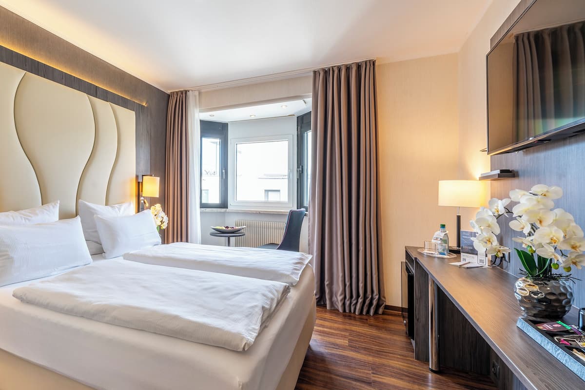 A hotel room with a large bed, two bedside tables with lamps, a desk with a computer and a vase of flowers, and a TV mounted on the wall.