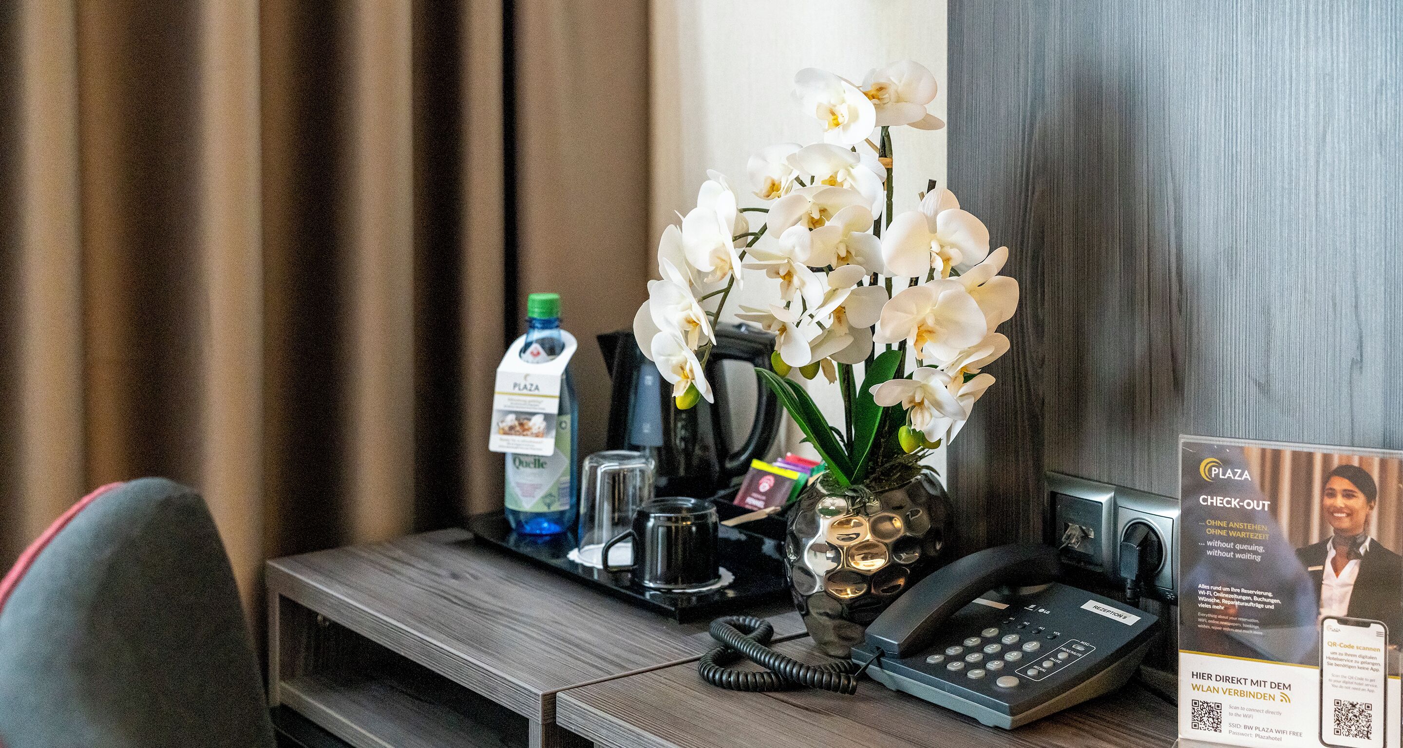 A hotel room desk with a telephone, a vase of white flowers, a bottle of water, and a coffee maker.
