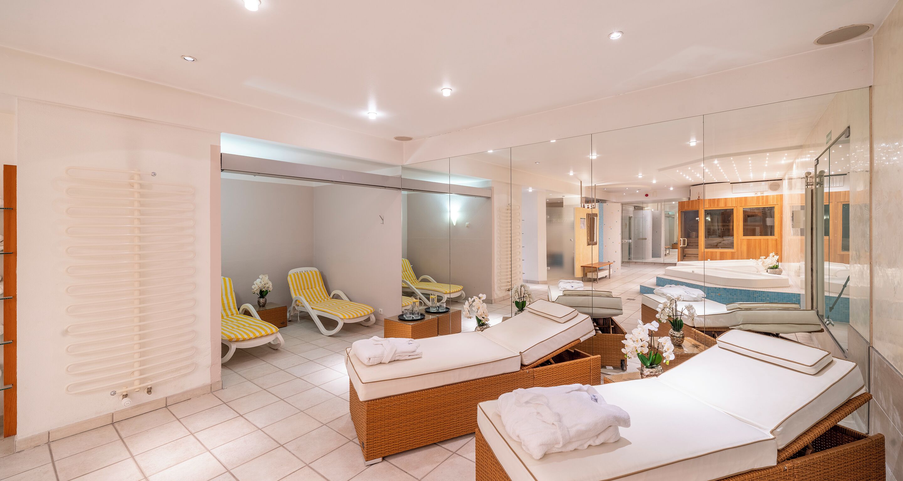 Un moderno cuarto de spa con un espejo grande, dos camastros, una camilla de masaje y una pequeña zona de estar con un banco y una mesa.