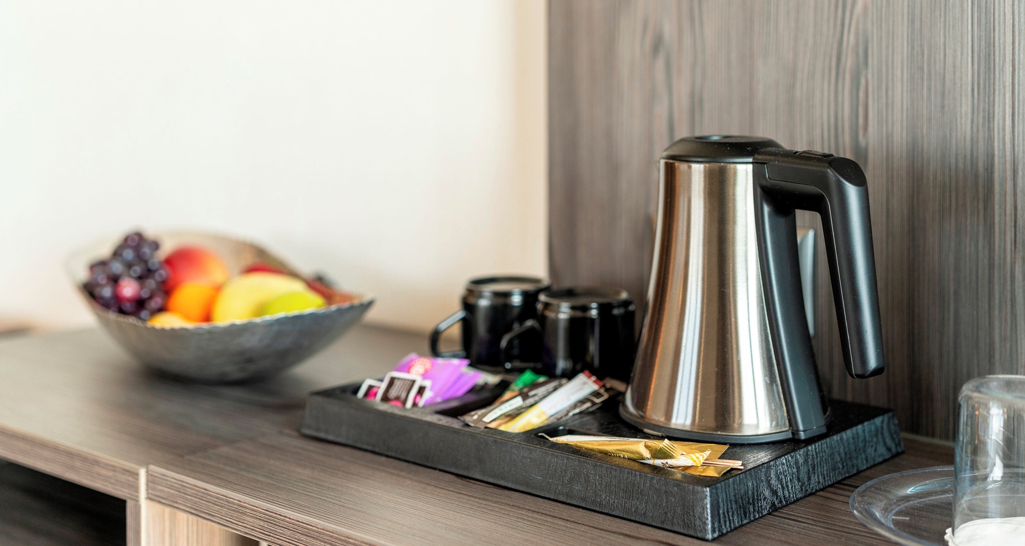 A stainless steel coffee maker on a black tray with a bowl of fruit and tea bags on a wooden surface.