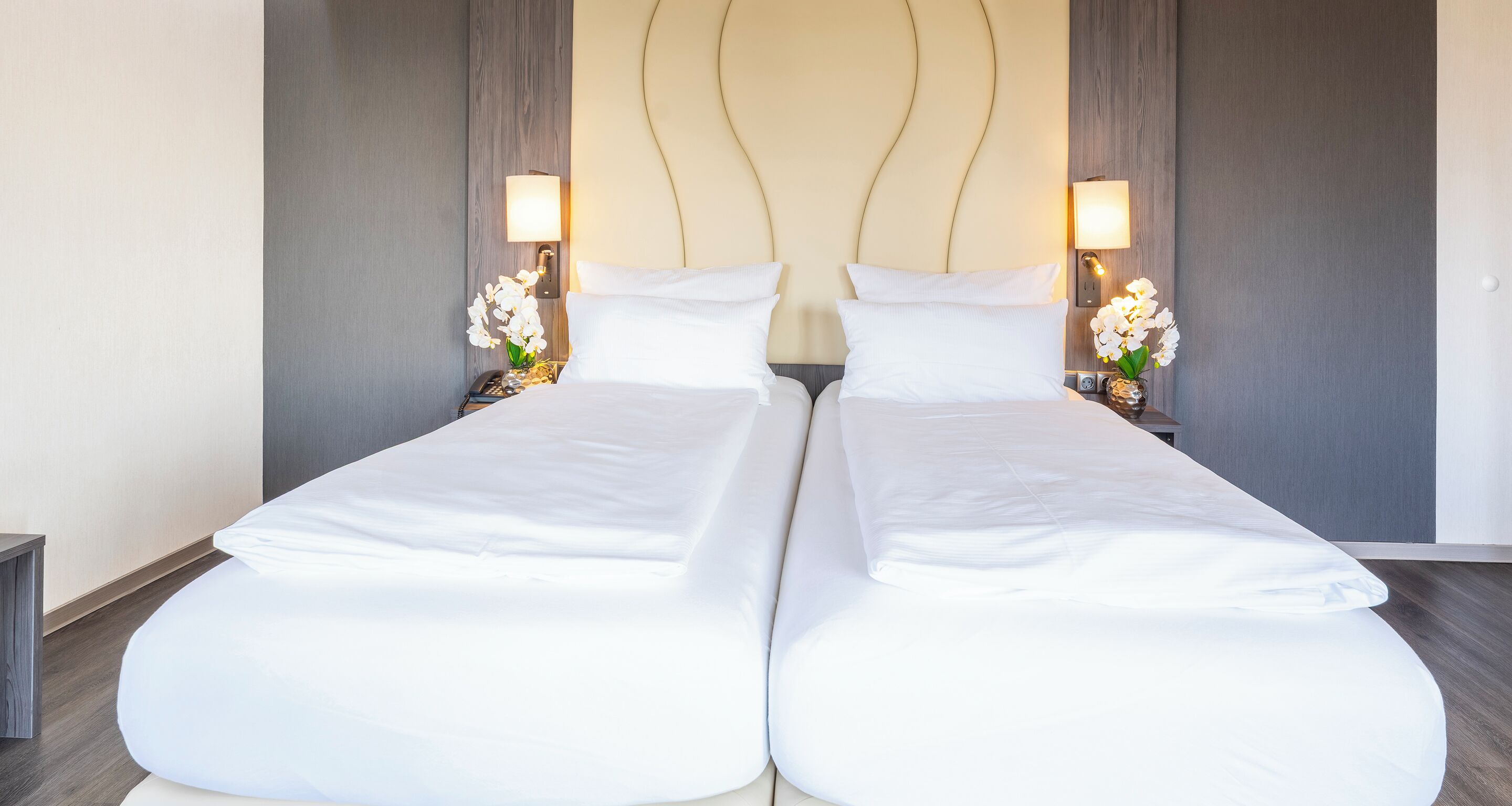 Una cama doble con sábanas blancas, dos mesitas de noche con lámparas y orquídeas blancas.