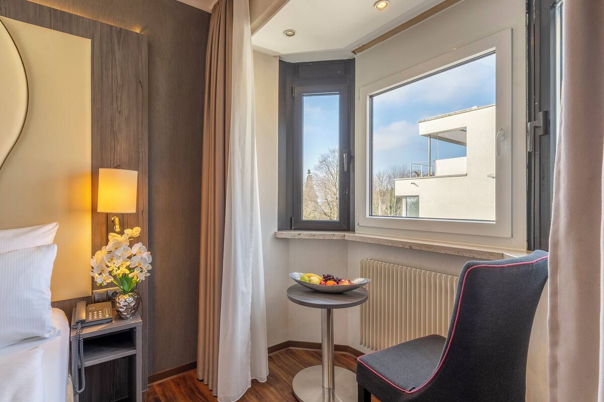 A hotel room with a bed, a bedside table with a lamp and phone, a chair, a small table with fruit, and a window with a view of buildings.