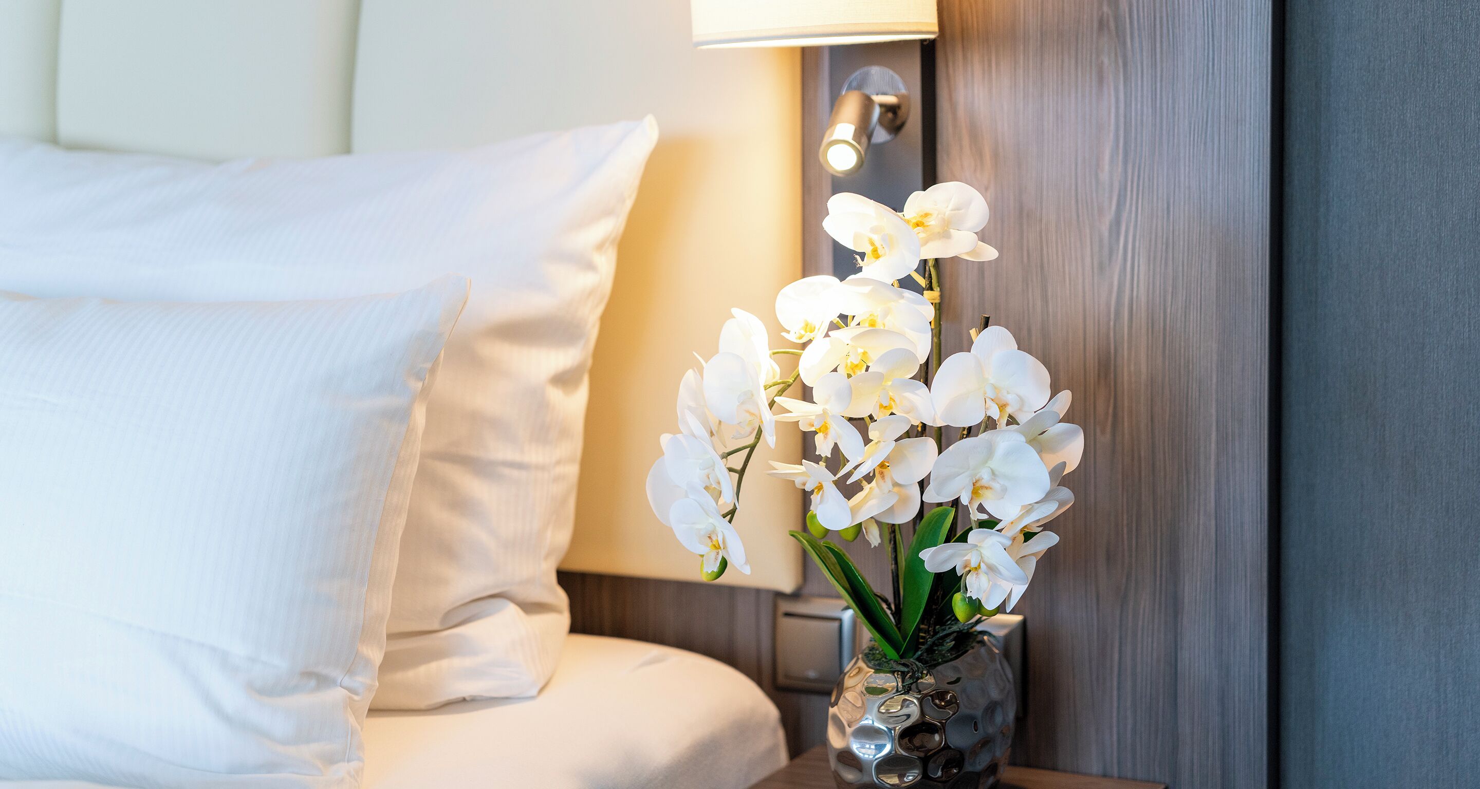 Ein Bett mit weißer Bettwäsche und eine Vase mit weißen Orchideen auf einem Nachttisch.