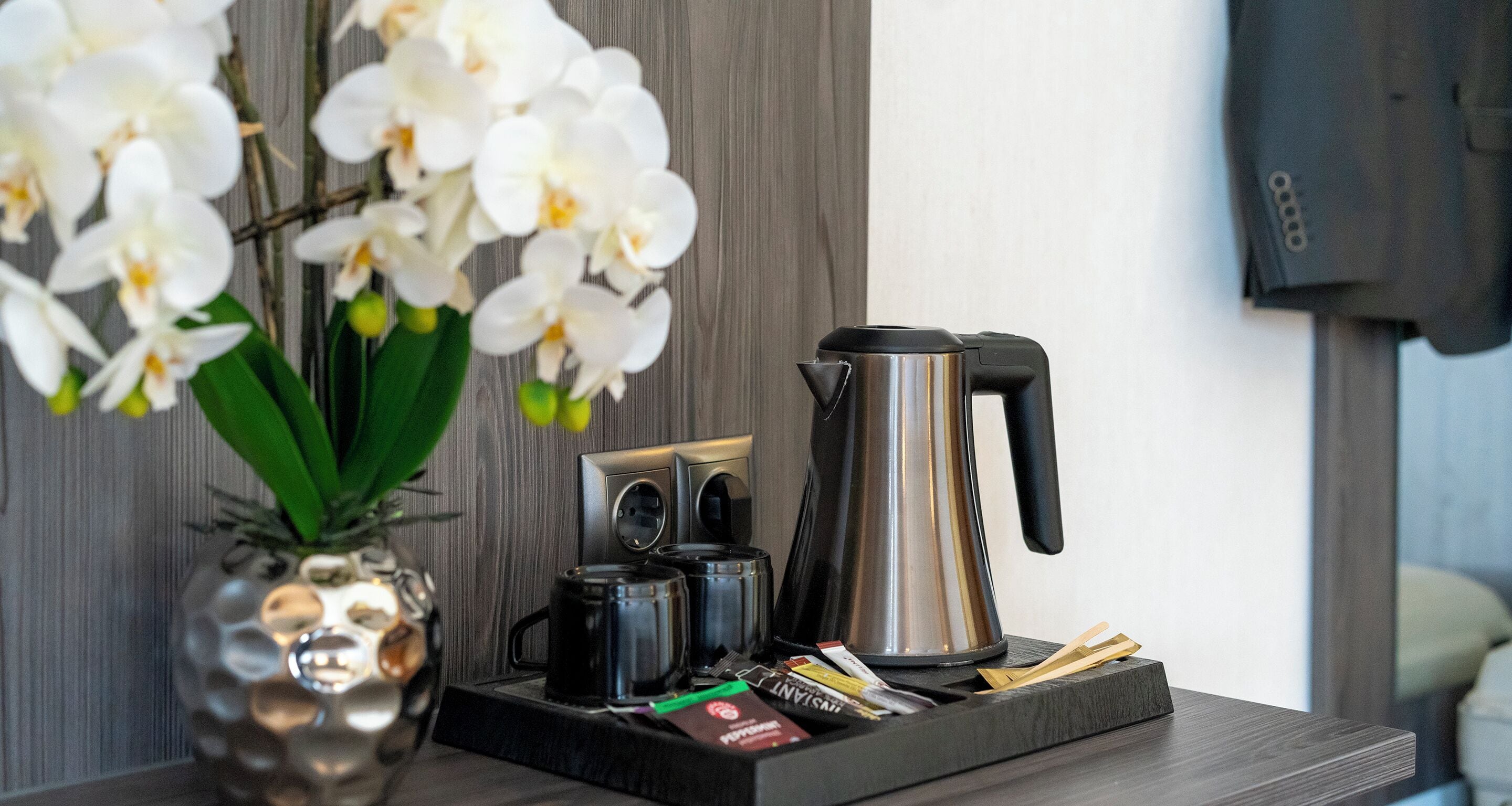 A coffee station with a stainless steel coffee pot, two black mugs, a small tray with coffee packets, and a decorative orchid vase.