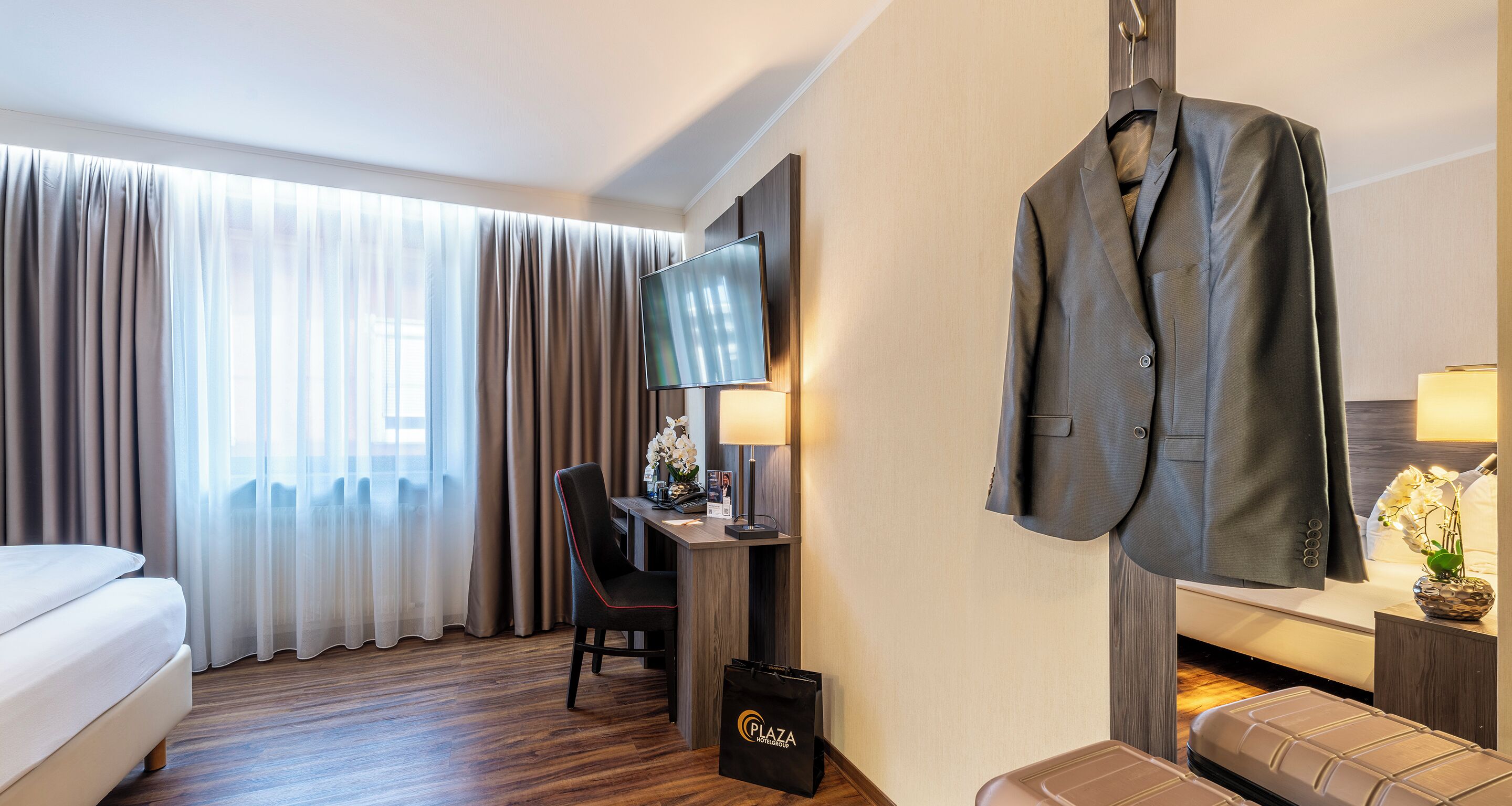 Hotelkamer met een bed, een bureau met stoel, een televisie, een kapstok met jas en een tas op de vloer.