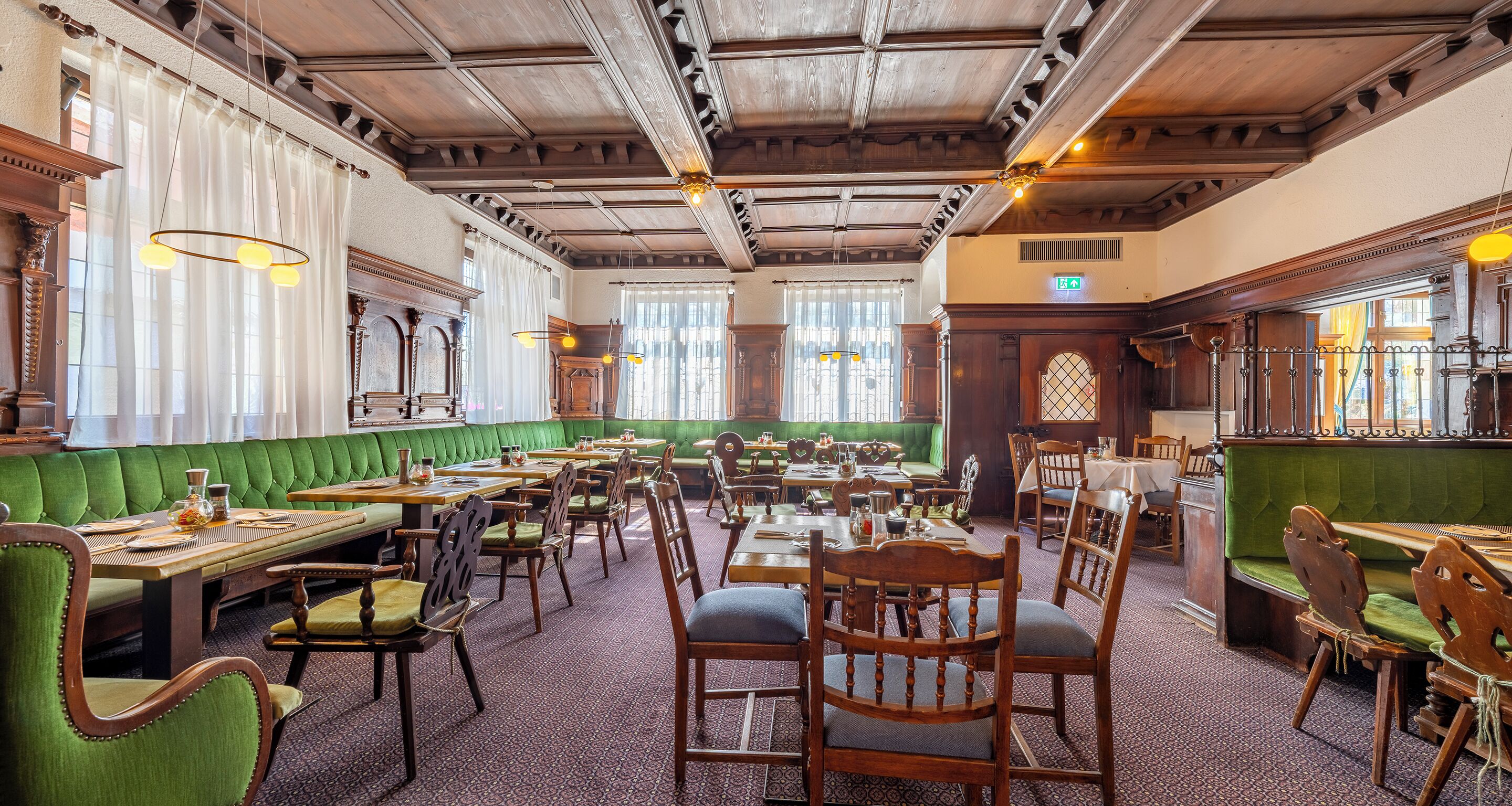 Un espace de restauration traditionnel, avec des meubles en bois, des banquettes recouvertes d’un tissu vert et des lustres classiques.