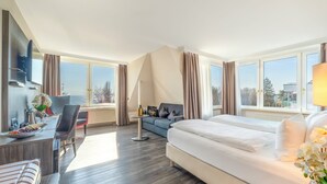 A hotel room with a bed, a sofa, a dining table with chairs, a TV, and large windows with curtains.