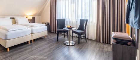 A hotel room with a bed, two armchairs, a small table with a bowl of fruit, a suitcase on a wooden side table, and a coat hanging on the wall.