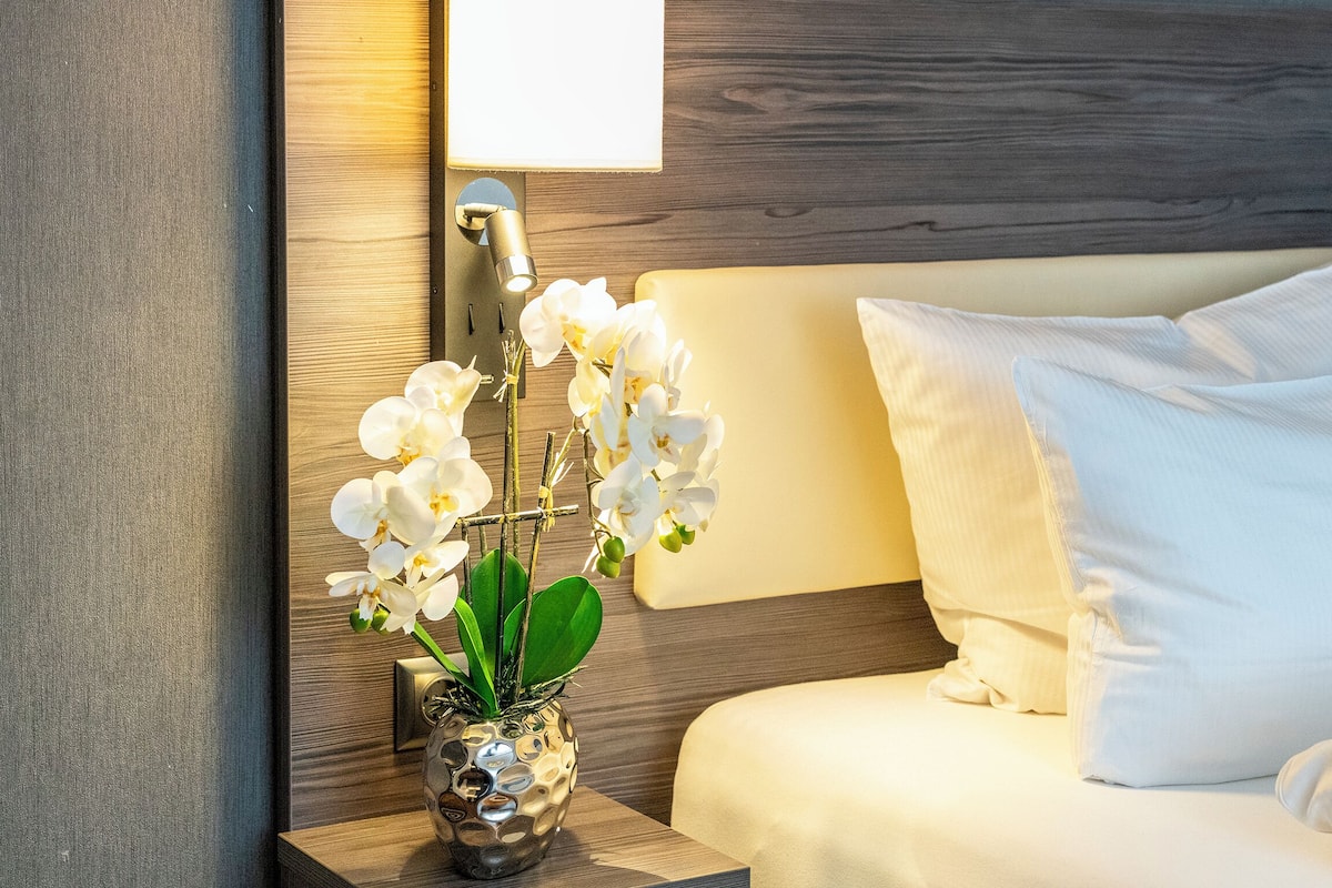 A neatly made bed with white pillows and a nightstand with a vase of white orchids.