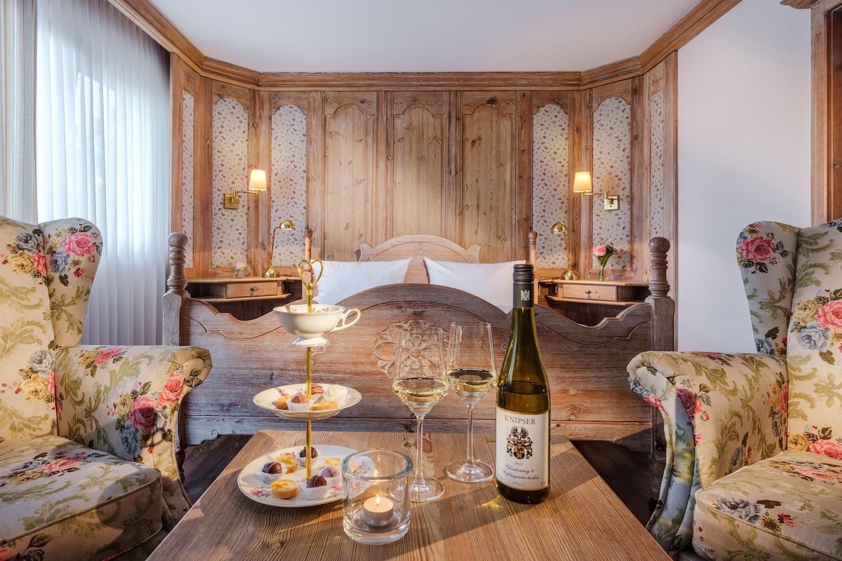 A cozy room with a wooden headboard, floral armchairs, a bedside table with a bottle of wine, glasses, and a tray of pastries.