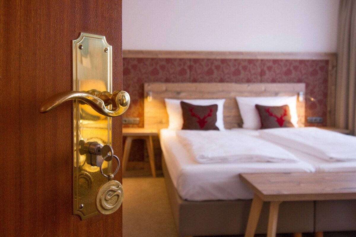 A hotel room with a wooden door, a bed with white bedding, and a nightstand with a lamp.