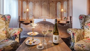 Una camera con una testiera in legno, poltrone floreali, un comodino con una bottiglia di vino, bicchieri e un vassoio di pasticcini.