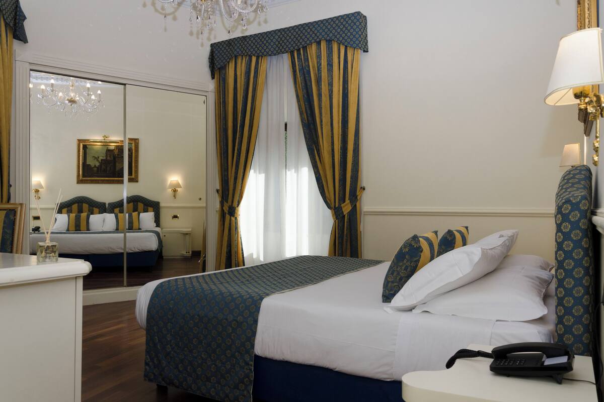Camera d'albergo con un letto grande, un comodino con lampada, un telefono e uno specchio.
