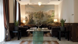 Un hall d’entrée avec un sol à carreaux, une table basse en verre, deux canapés et une grande fresque représentant un paysage fluvial.