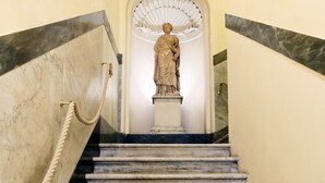 Un escalier en marbre, surmonté d’une statue, mène à une pièce dotée d’un plafond blanc et d’un éventail décoratif.
