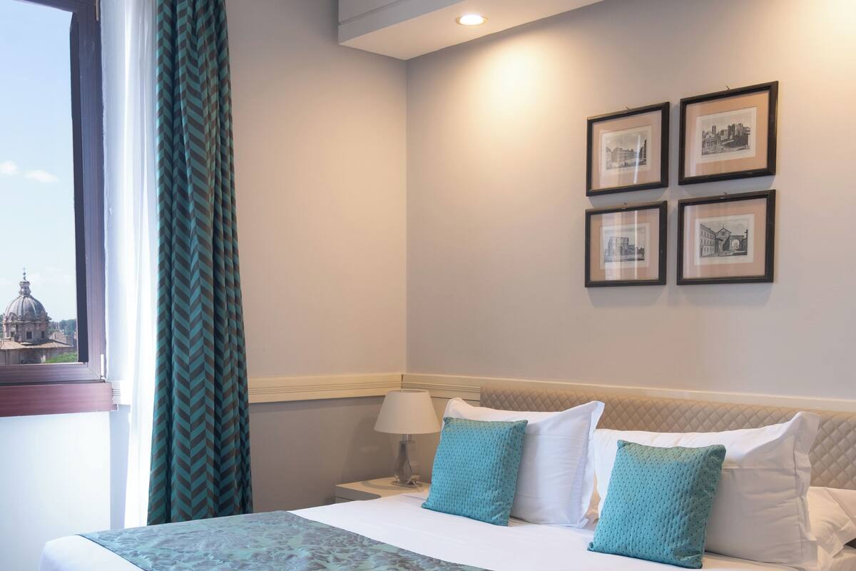 Un letto rifatto con una coperta a motivi, una lampada da comodino e una finestra con vista su un edificio.