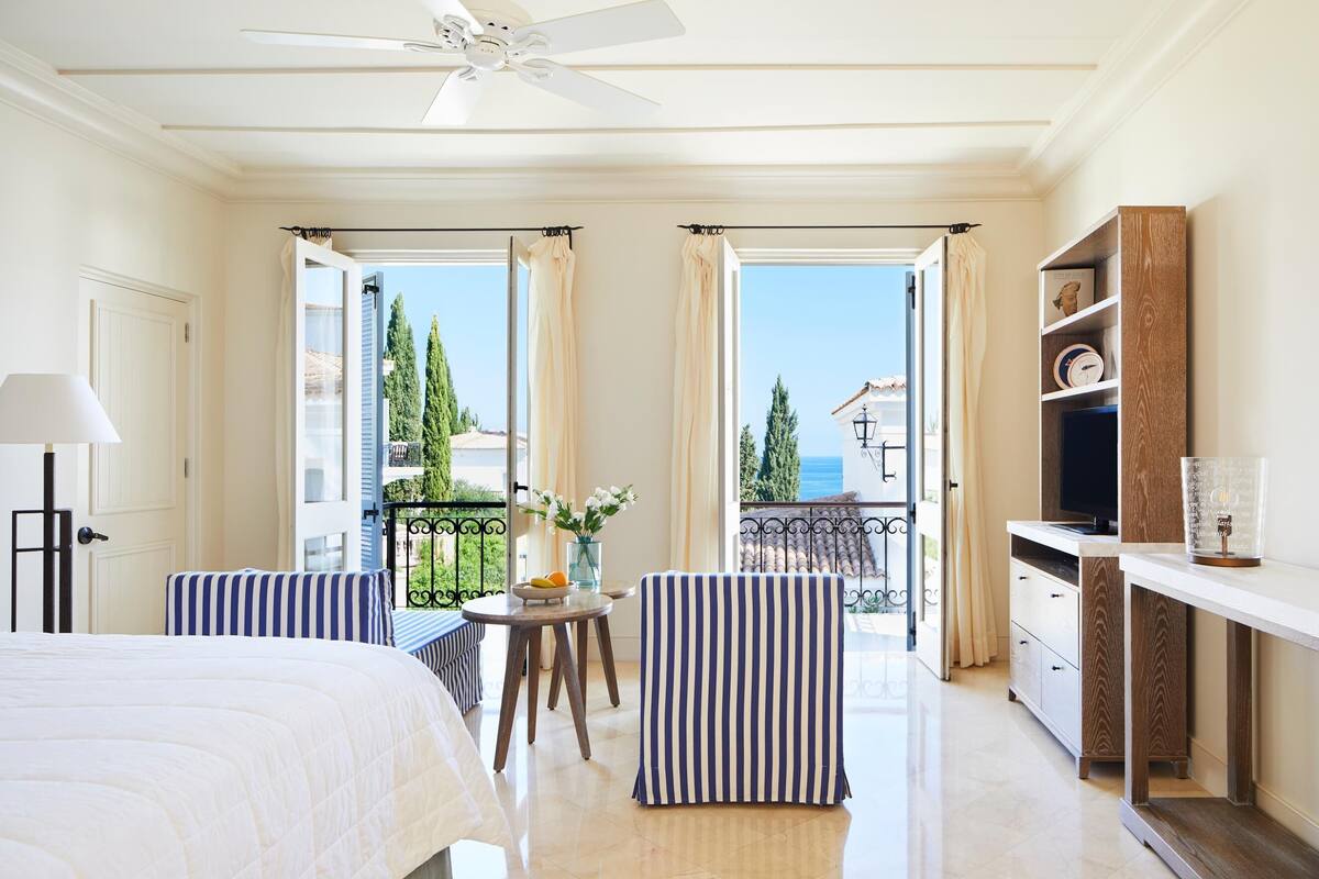 A hotel room with a bed, a desk, a chair, a small table with flowers, and a view of the sea.