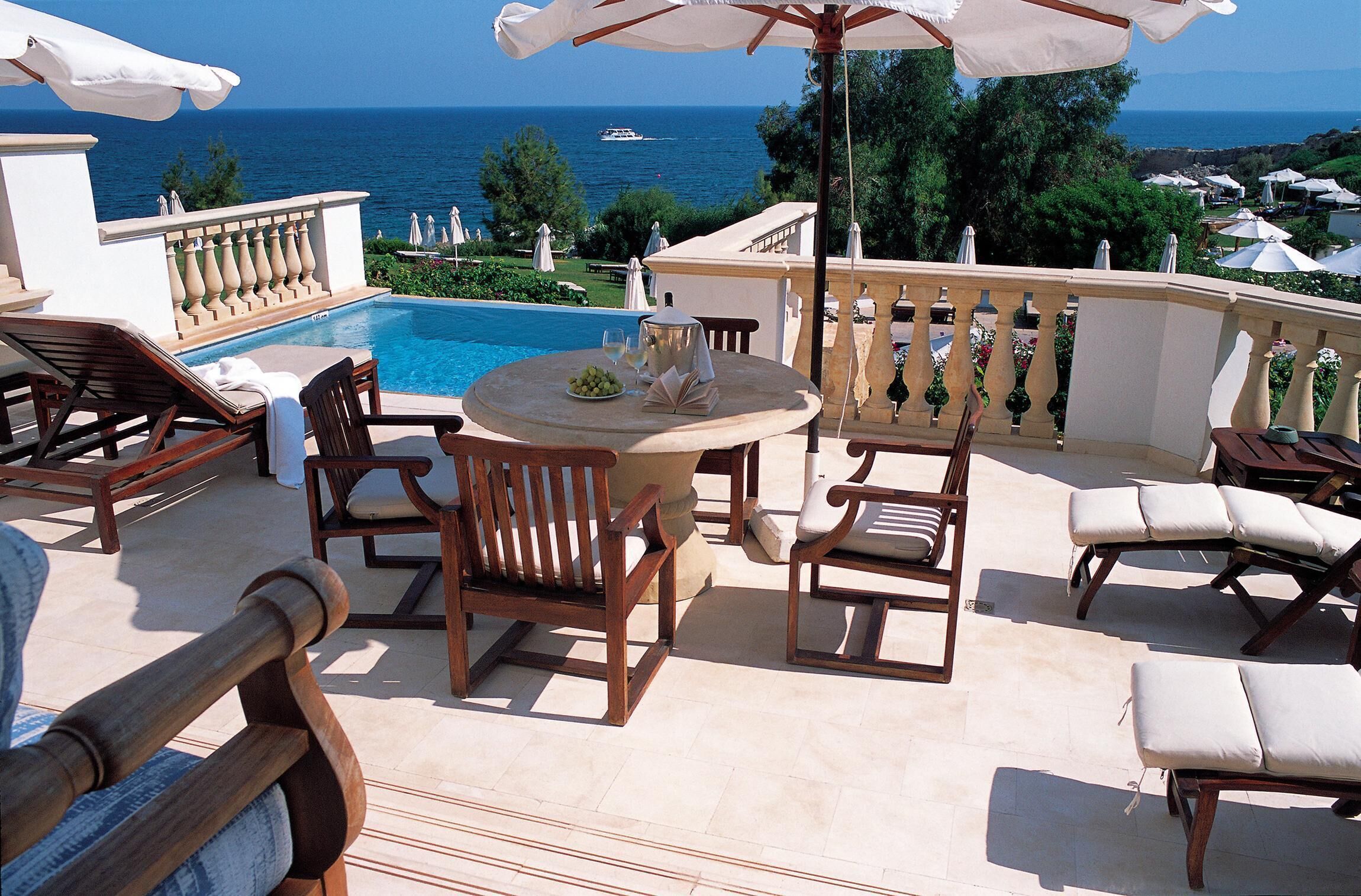 A poolside area with lounge chairs, a table with a bowl of fruit and a glass of wine, and a view of the sea.