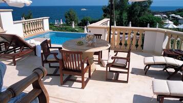 A poolside area with lounge chairs, a table with a bowl of fruit and a glass of wine, and a view of the sea.
