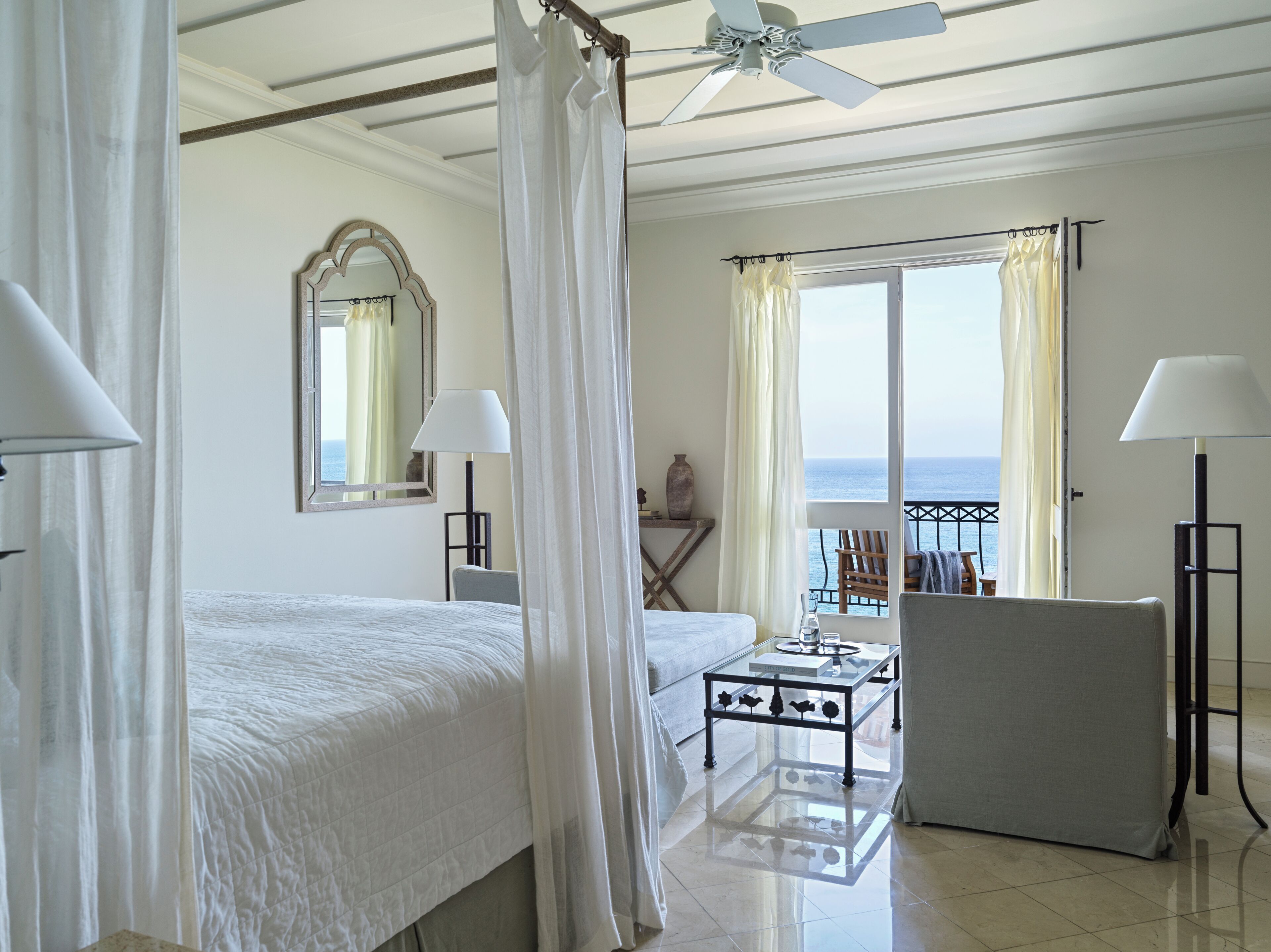A hotel room with a bed, a desk, a chair, a balcony with a view of the sea, and a ceiling fan.