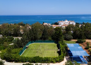A tennis court surrounded by trees and buildings, with a view of the sea.