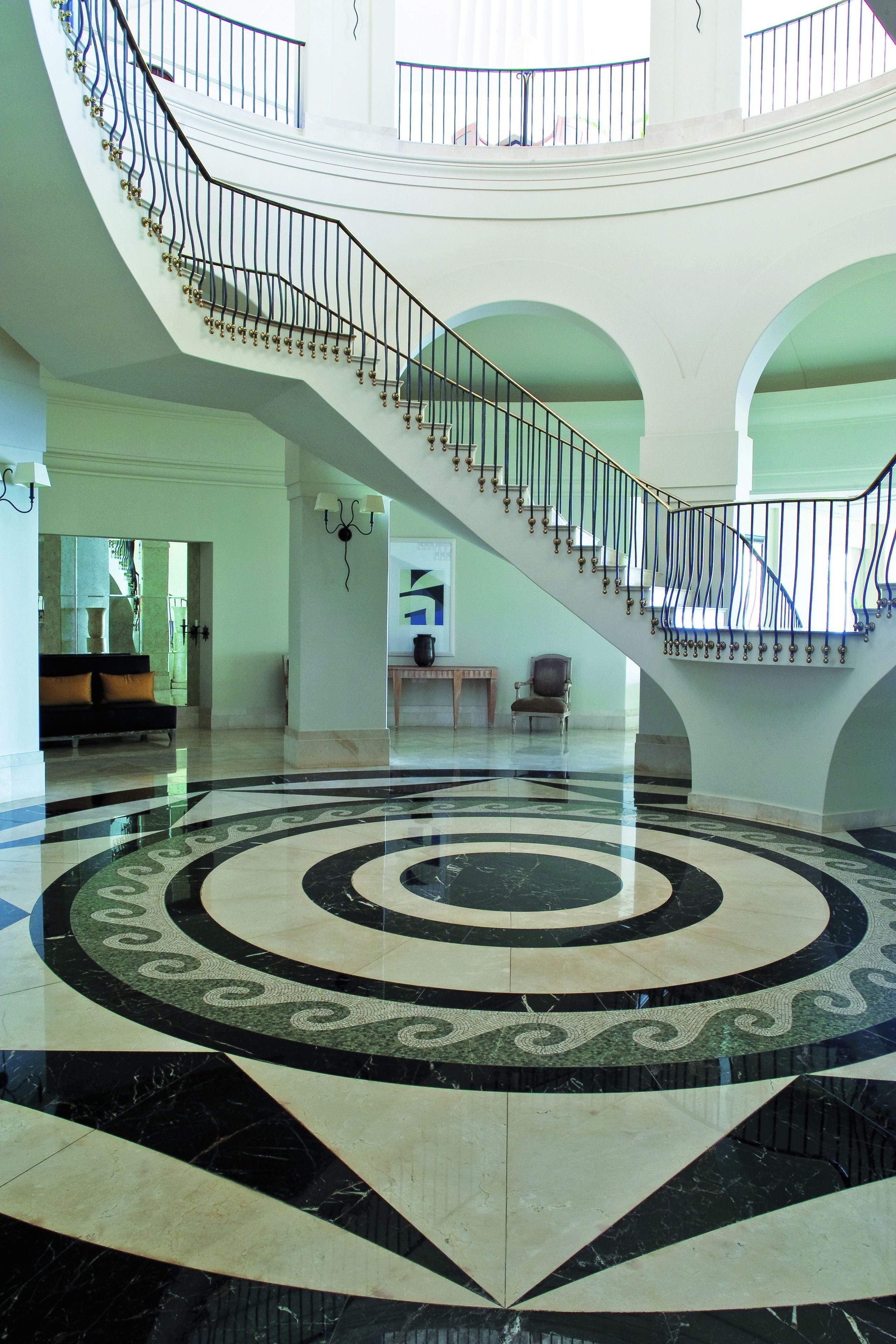 A grand lobby with a central circular patterned floor, a staircase with a black railing, and a seating area with a sofa and chairs.
