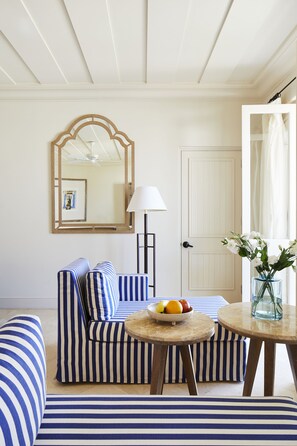 A room with striped blue and white furniture, a round table with a fruit bowl, a mirror, a lamp, and a door.