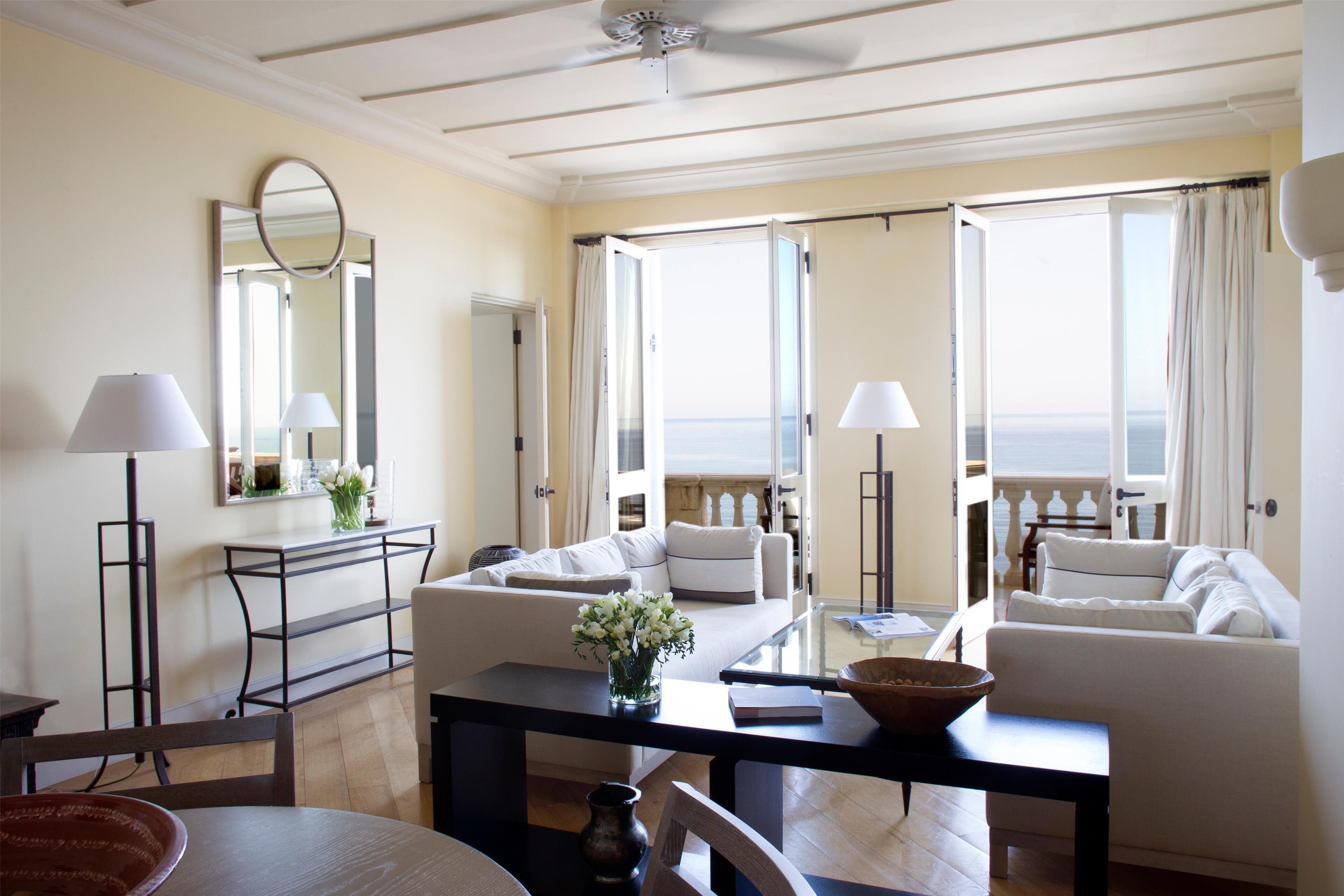 A spacious living room with a large window overlooking the sea, a white sofa, a coffee table, and a ceiling fan.