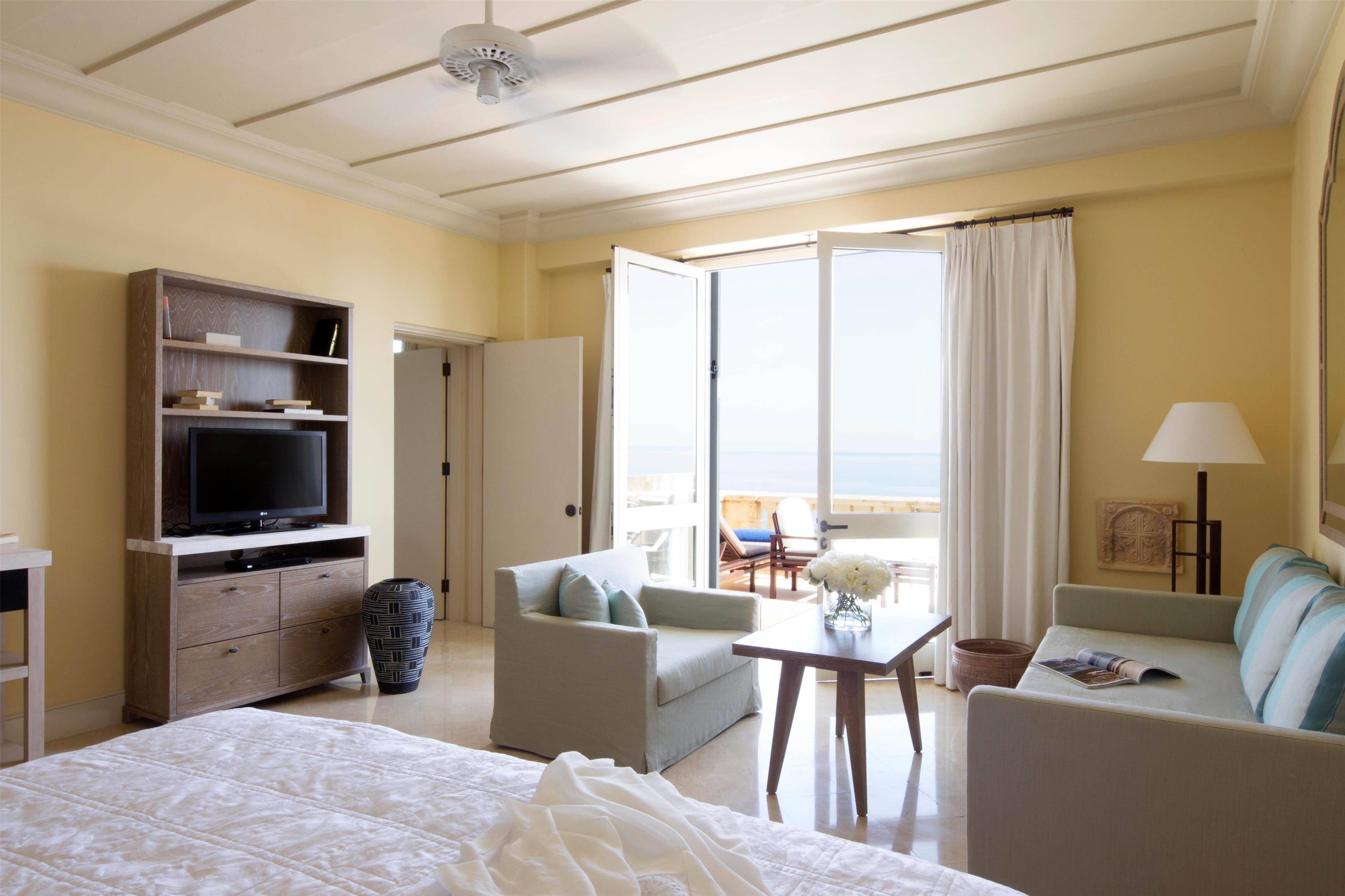 Una camera d'hotel con un letto, una televisione, un divano, una sedia, un tavolo e vista sul mare attraverso un'ampia finestra.